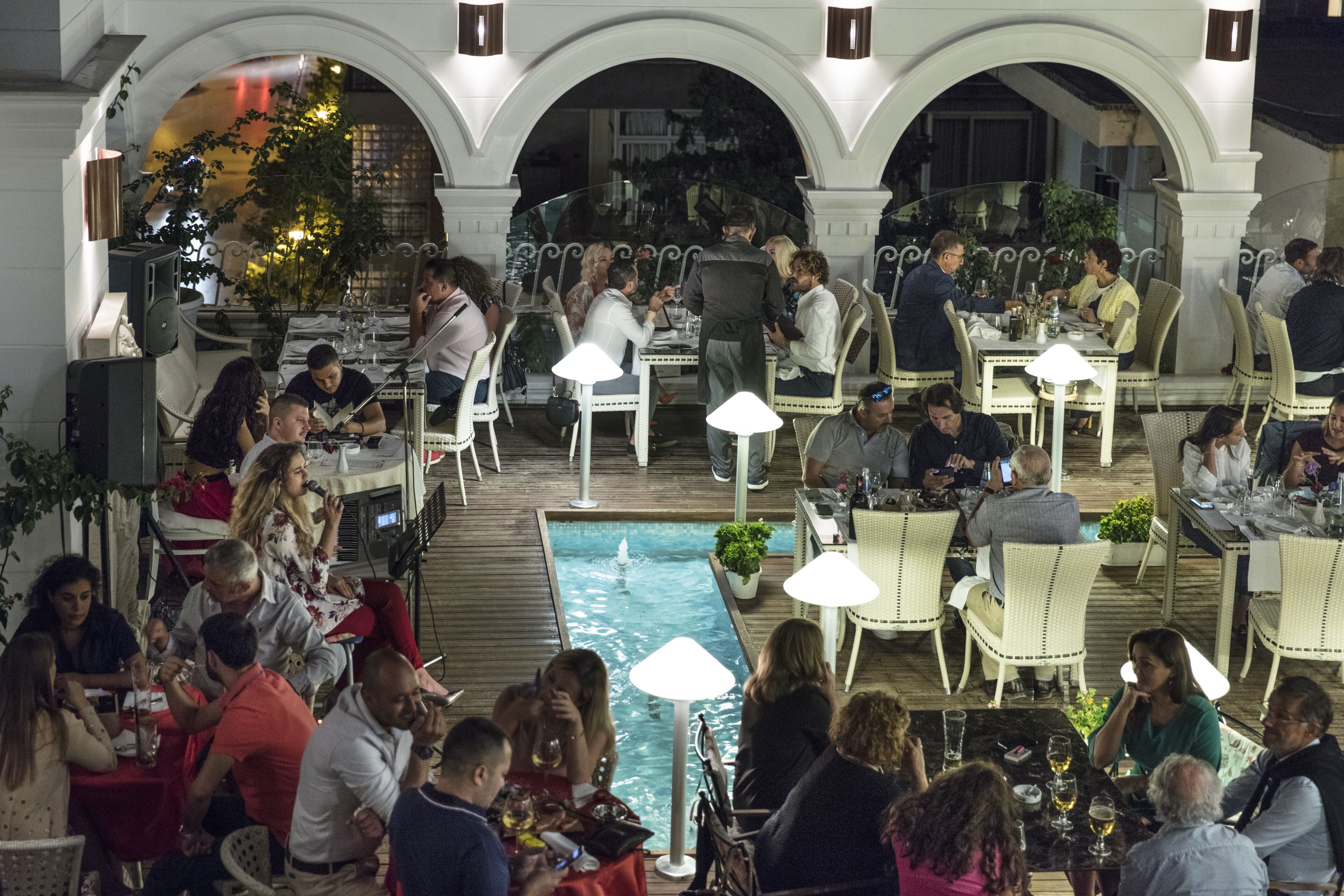 An outdoor dining area with multiple tables and chairs, a central pool, and an arched architectural design.