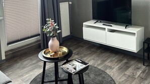 Een moderne woonkamer met een flatscreen televisie op een witte tv-meubel, een klein rond tafeltje met een vaas en een dienblad, en een grijs tapijt.