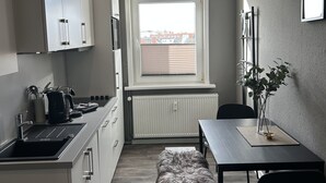 Een kleine keuken met witte kastjes, een zwart aanrechtblad en een raam met zonwering.