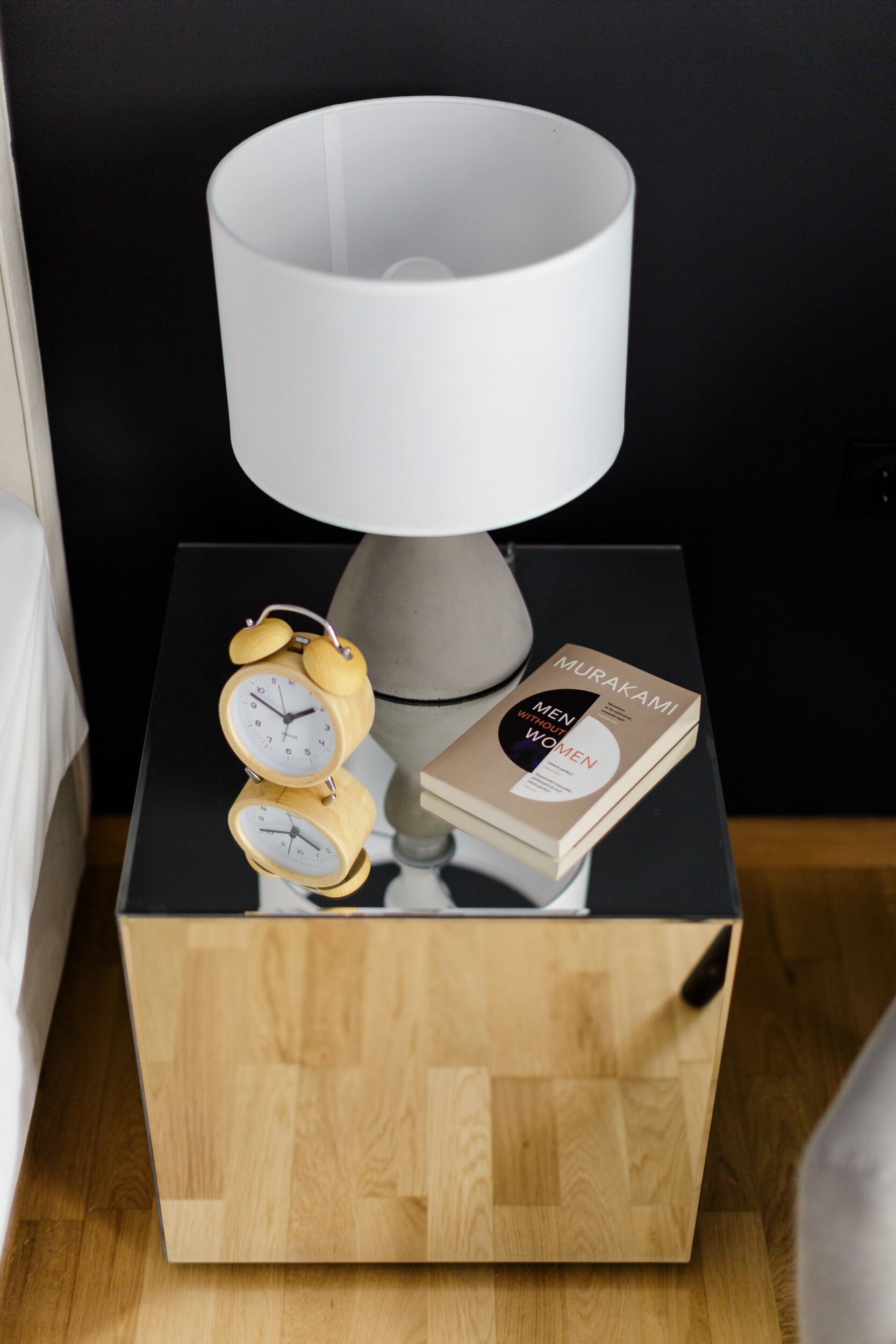 A bedside table with a wooden side table, a white lamp, two wooden alarm clocks, and a book titled "Men Without Women" by Murakami.