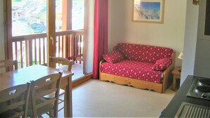 Ein gemütliches Zimmer mit einem Holzespeisetisch und Stühlen, einem roten, gemusterten Sofa, einem kleinen Holzschemel und einem Bild an der Wand.