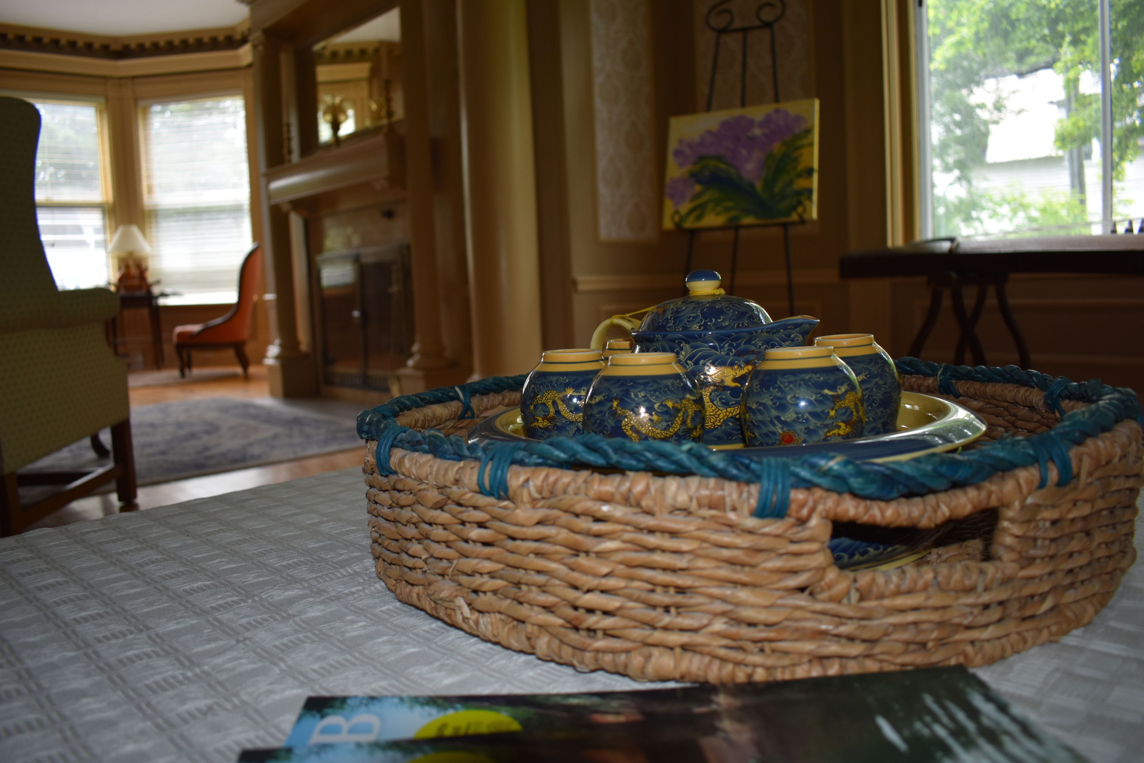 Een rieten mand met blauwe en gouden decoratieve objecten op een tafel in een kamer met een open haard, een schilderij en uitzicht door een raam.