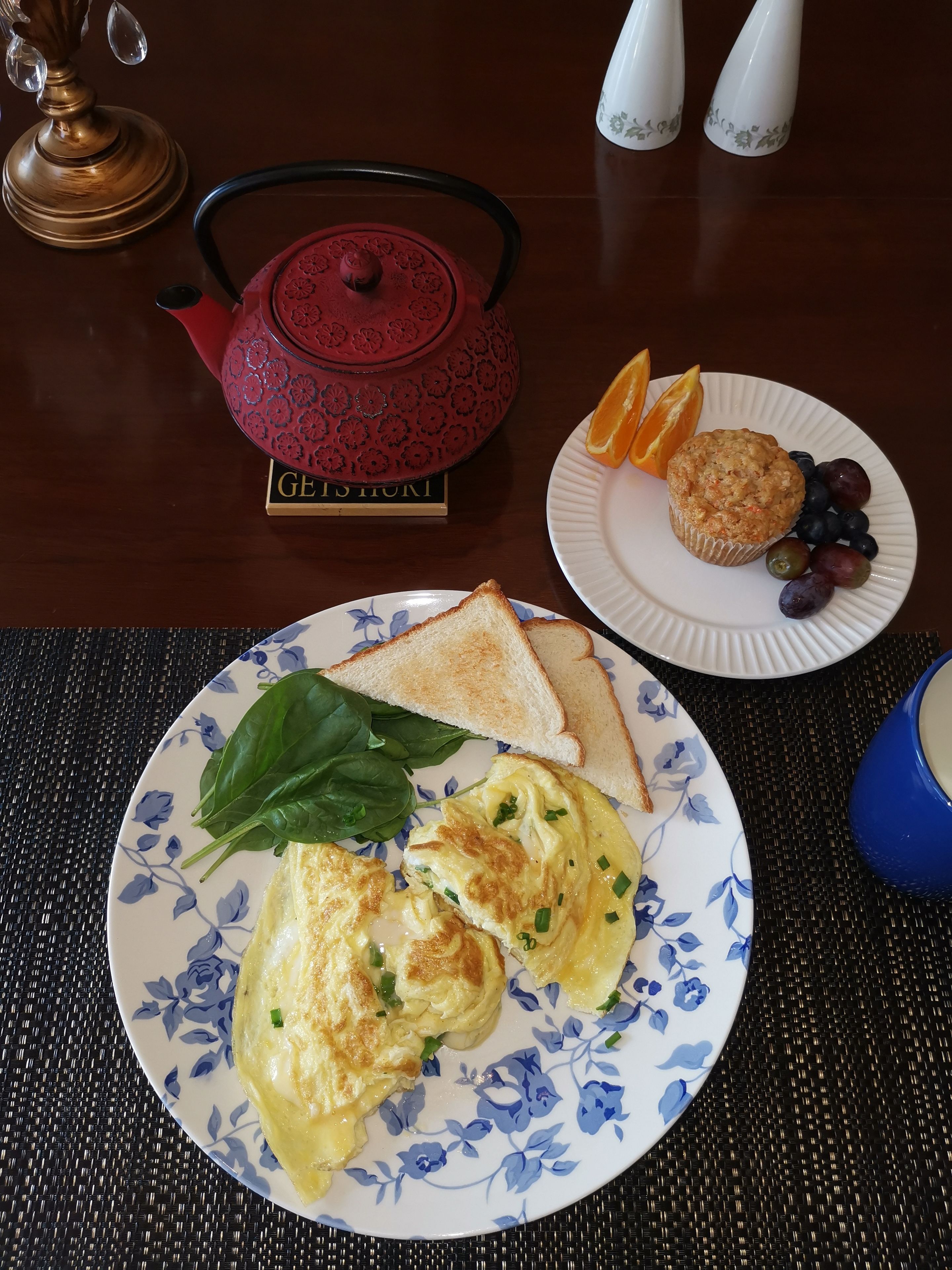 A breakfast setting with toast, spinach, scrambled eggs, a muffin, and fruit on plates, a teapot, and salt and pepper shakers on a table.