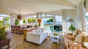 A spacious living room with a large window offering a scenic view, a dining area with a table and chairs, and a cozy seating area with sofas and a coffee table. - House Puerto Pollensa Llenaire with pool (Puerto Pollensa)