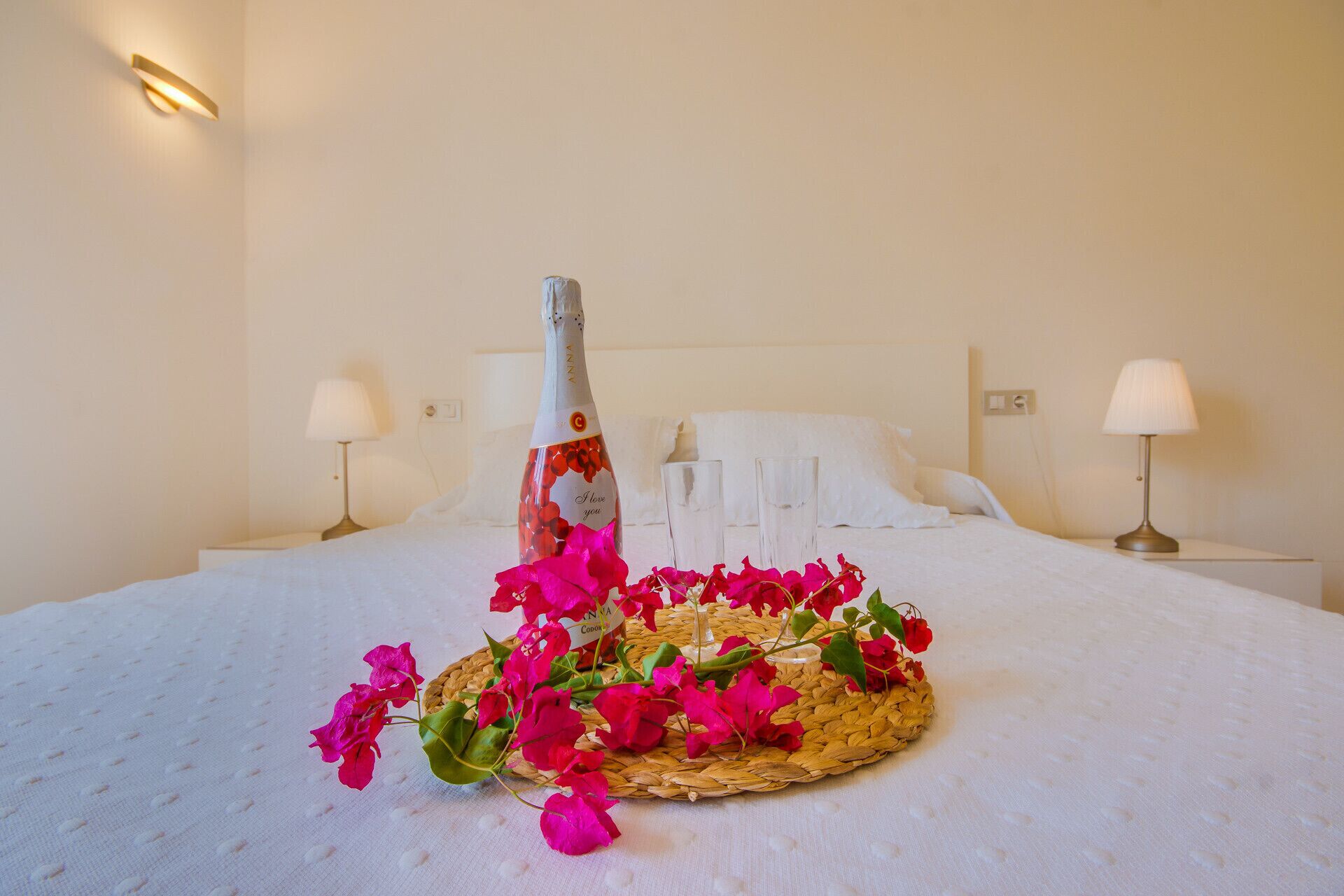 Een keurig opgemaakt bed met een fles champagne, een geweven mand met bloemen en twee bedlampjes in een hotelkamer.