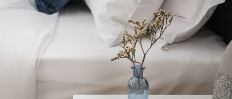A neatly made bed with white and blue bedding, a vase with dried flowers, and a lamp on a bedside table.