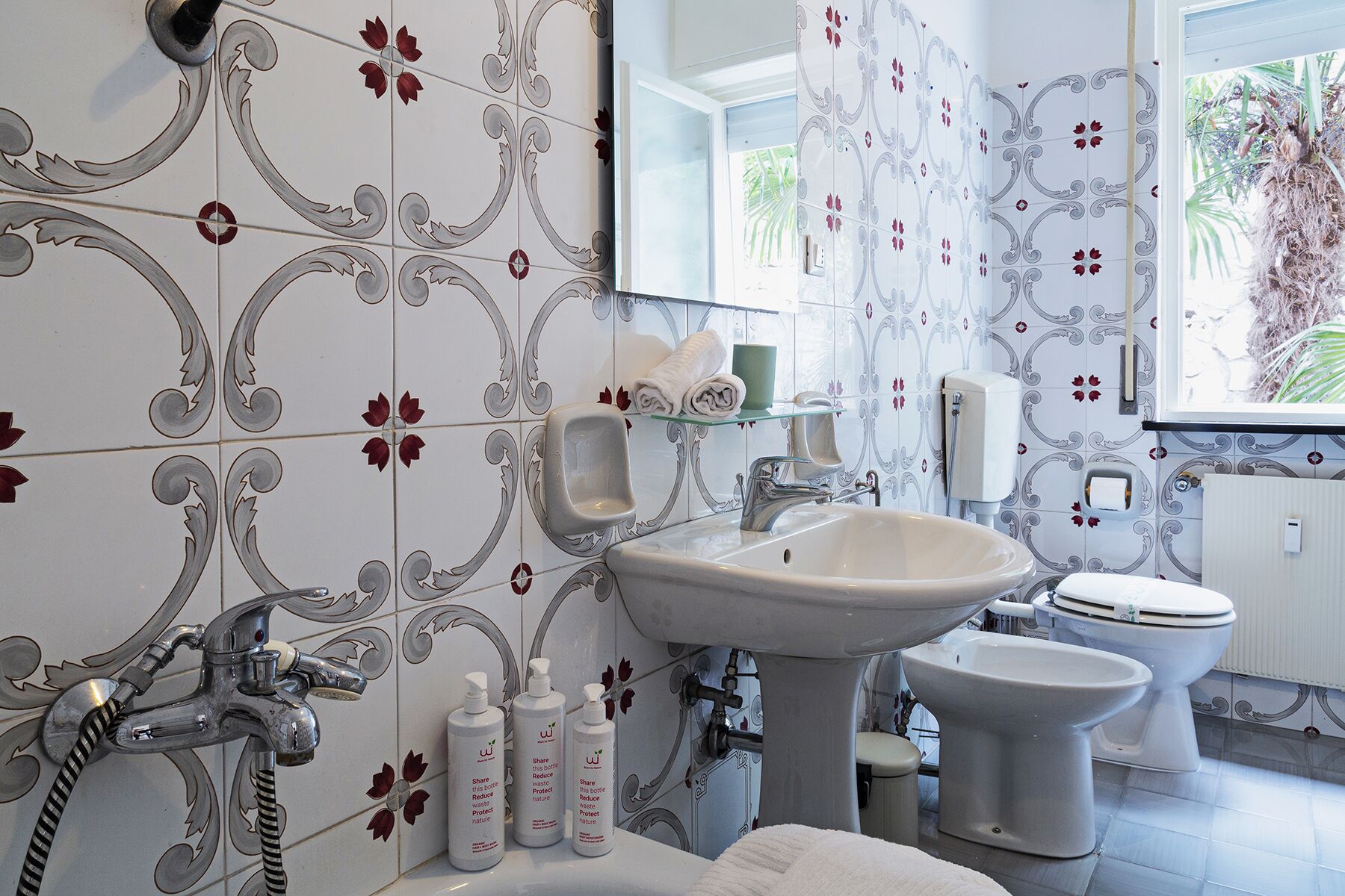 A bathroom with a large sink, a toilet, and a bidet. There are decorative tiles on the walls and a window with a view of palm trees.