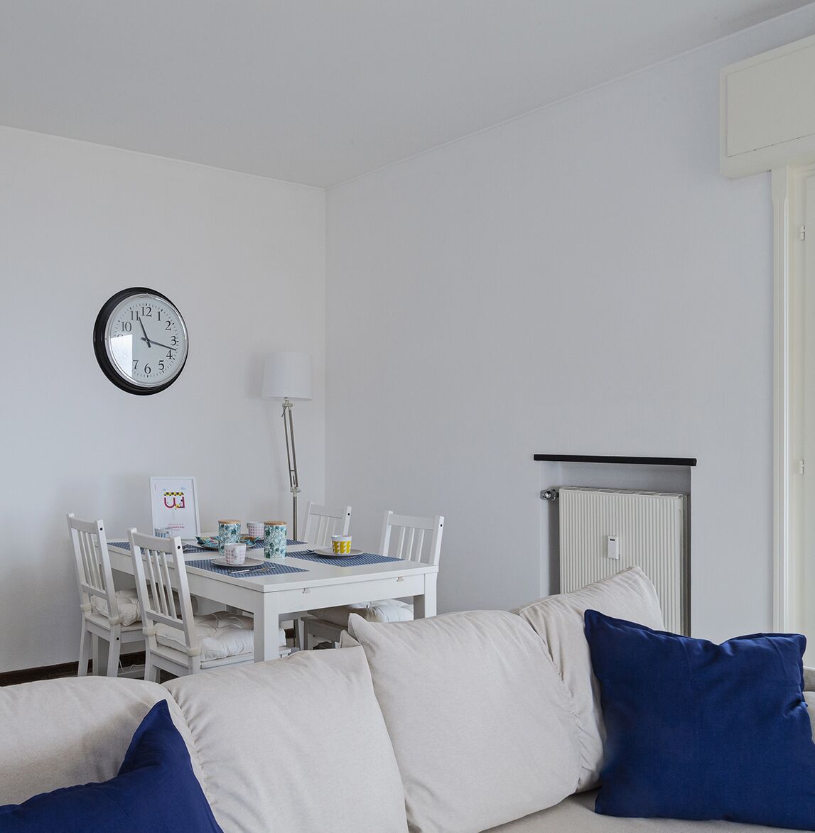 Ein Zimmer mit weißem Tisch, Stühlen, einer Uhr, einer Heizung und einer Couch mit blauen Kissen.