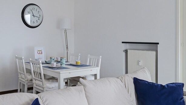 Ein Zimmer mit weißem Tisch, Stühlen, einer Uhr, einer Heizung und einer Couch mit blauen Kissen.