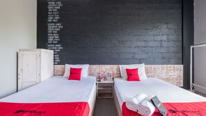 Une chambre d’hôtel avec deux lits, chacun doté d’un matelas blanc et de draps rouges. Une table de chevet en bois se trouve entre les lits, avec une bouteille d’eau et un téléphone. Le mur derrière les lits est noir avec du texte blanc.