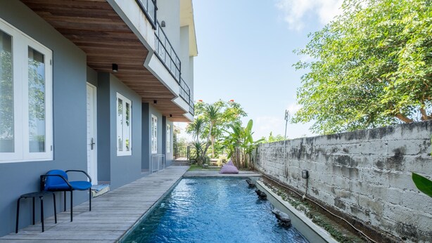 Une piscine dotée d’une terrasse en bois et de chaises bleues, entourée d’un mur en béton et de verdure.