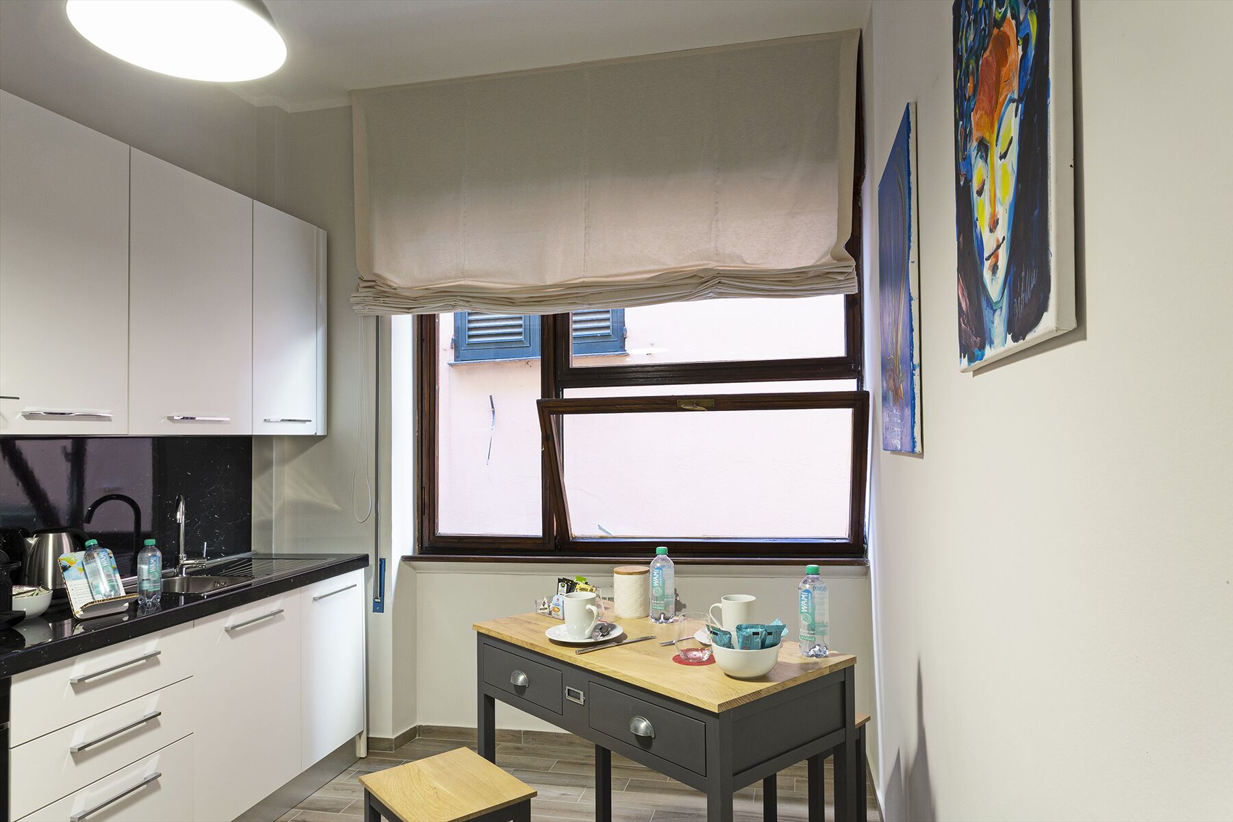 Una cocina compacta con armarios blancos, una encimera negra, una mesa de madera y un ventanal con un valance color beige.