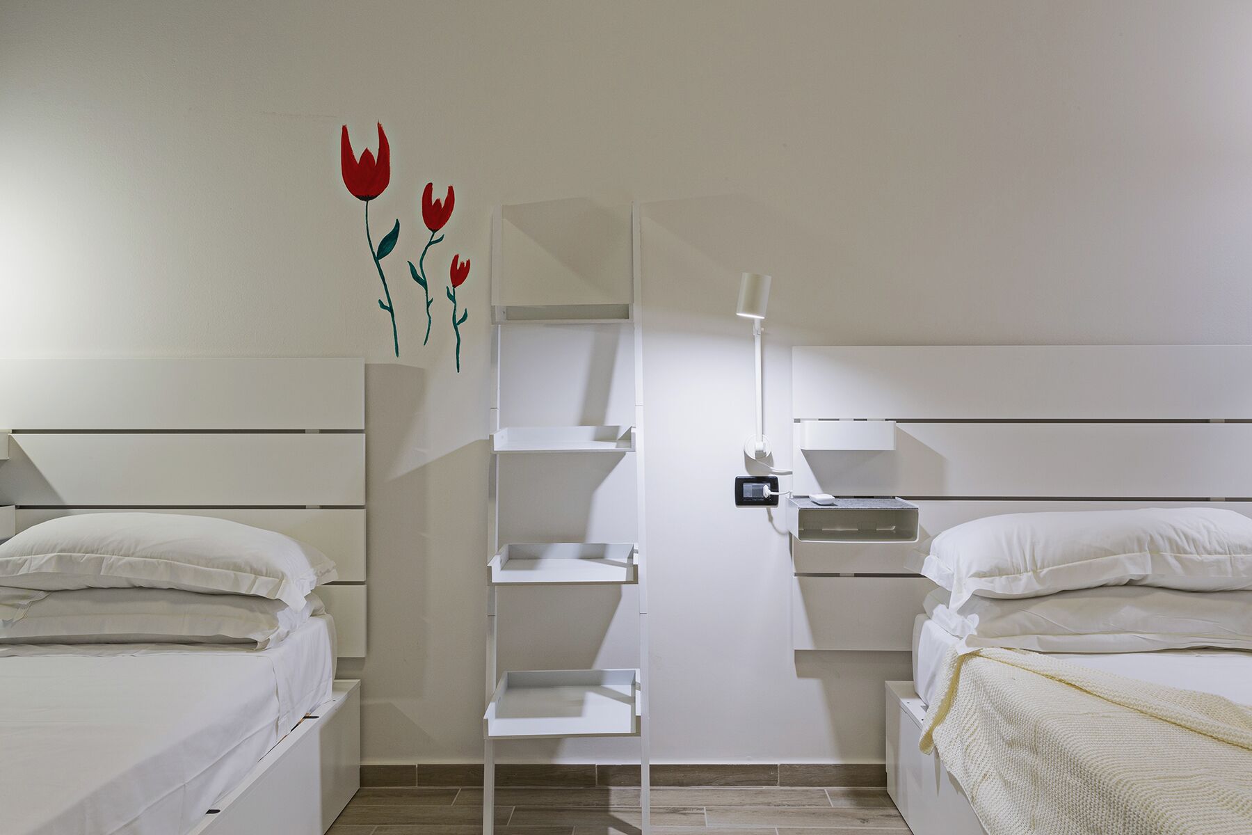 A hotel room with two beds, a white wardrobe, a shelf unit, and a bedside table with a lamp.