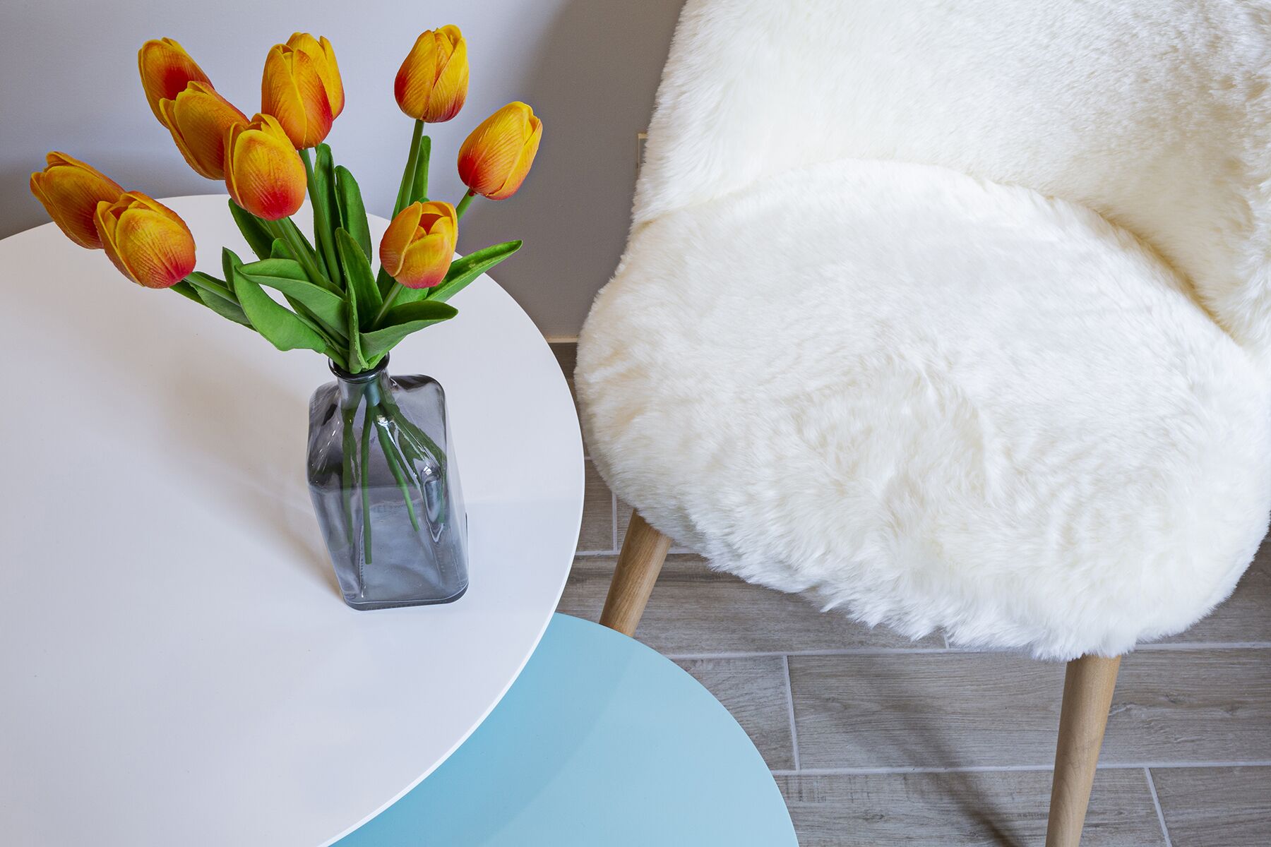 A white chair with a fluffy white cover, a clear glass vase with yellow tulips, and a round table with a blue and white design.