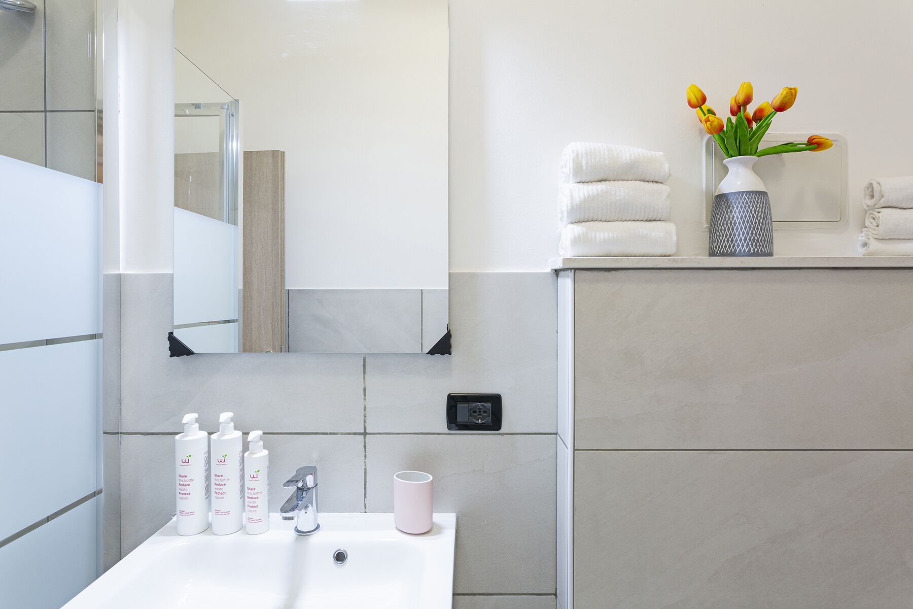 Ein modernes Badezimmer mit Waschbecken, Pflegeprodukten, Spiegel und einer Vase mit gelben Tulpen.
