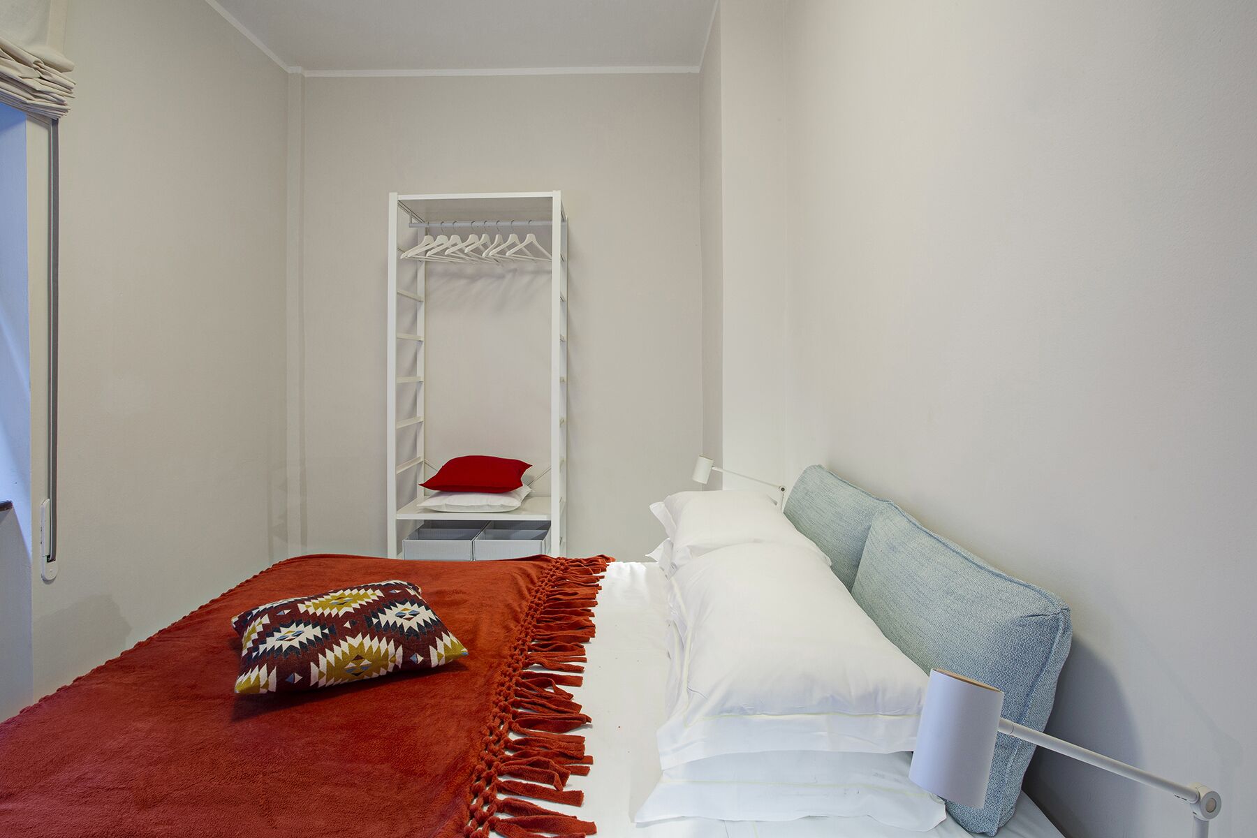 A small, neatly organized bedroom with a bed, a bedside table, a hanging shelf, and a wardrobe.