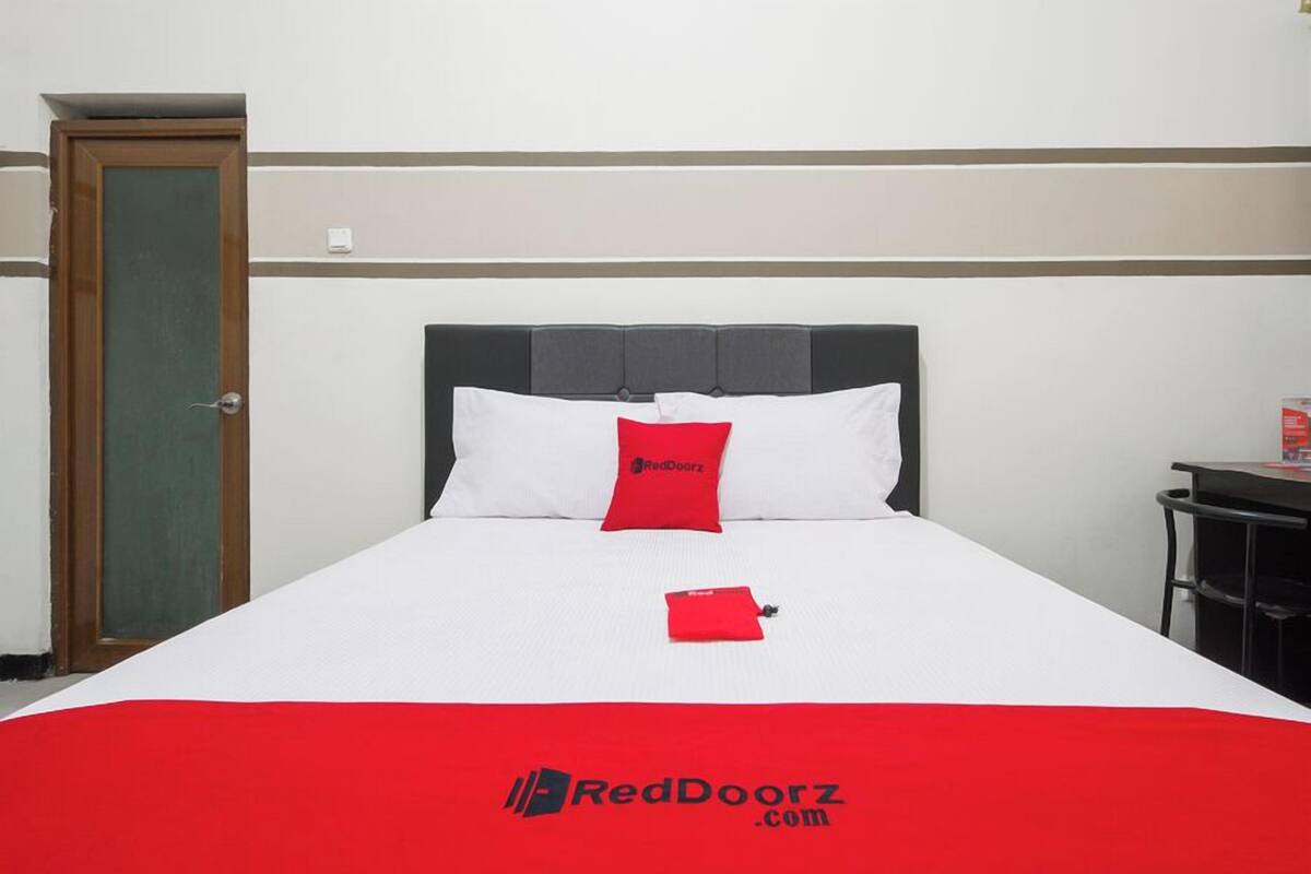 A hotel room with a bed, a red pillow, a red blanket, a door, and a small table with a red object on it.