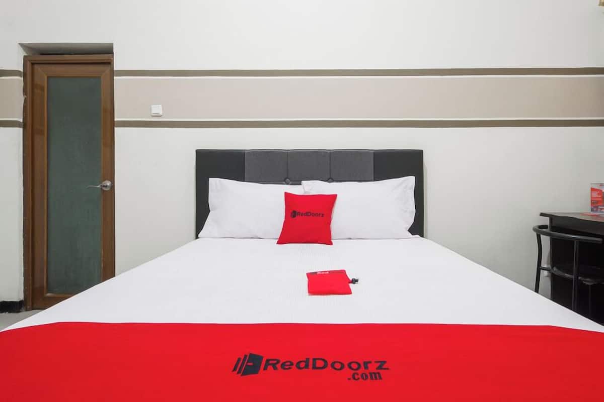 Habitación de hotel con cama, almohada roja, colcha roja, una puerta y una pequeña mesa con un objeto rojo encima.