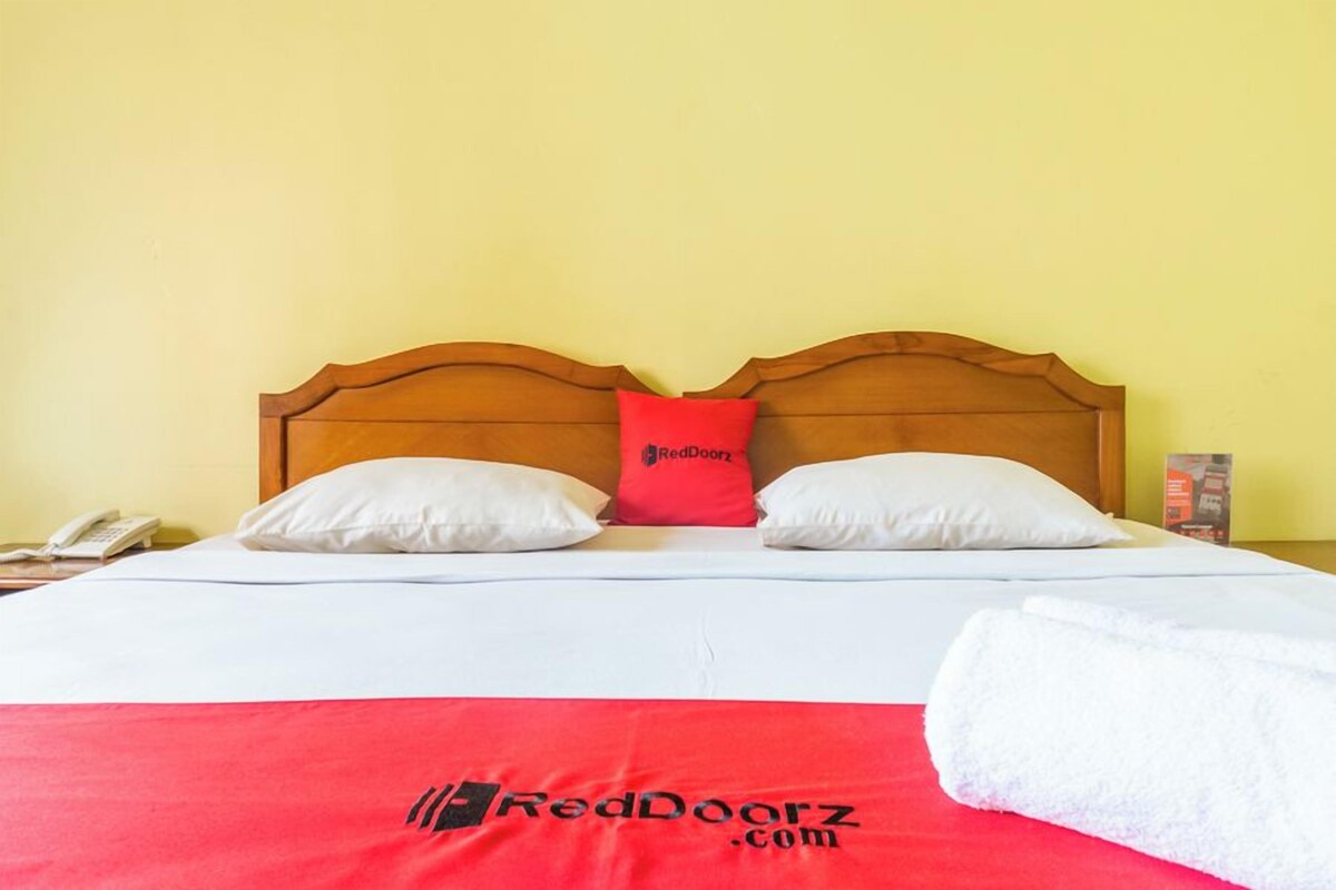 A bed with a wooden headboard, two white pillows, a red pillow, a white blanket, and a rolled white towel.
