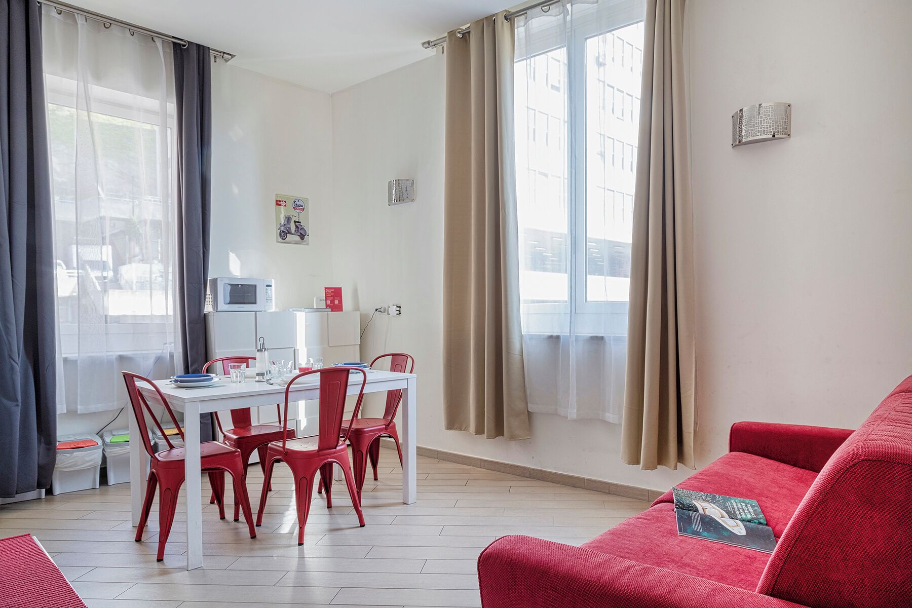A modern living room with a red sofa, a dining table with red chairs, a white microwave, and a window with beige curtains.