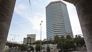 Um edifício alto e moderno com fachada azul e branca, cercado por árvores e uma rua com semáforos e placas de trânsito.