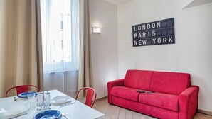 Quarto com mesa de jantar posta para dois, um sofá vermelho e um letreiro na parede listando Londres, Paris e Nova Iorque.