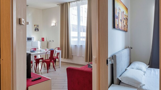 Quarto de hotel com cama, escrivaninha com cadeira, pequeno espaço de cozinha e janela com cortinas.