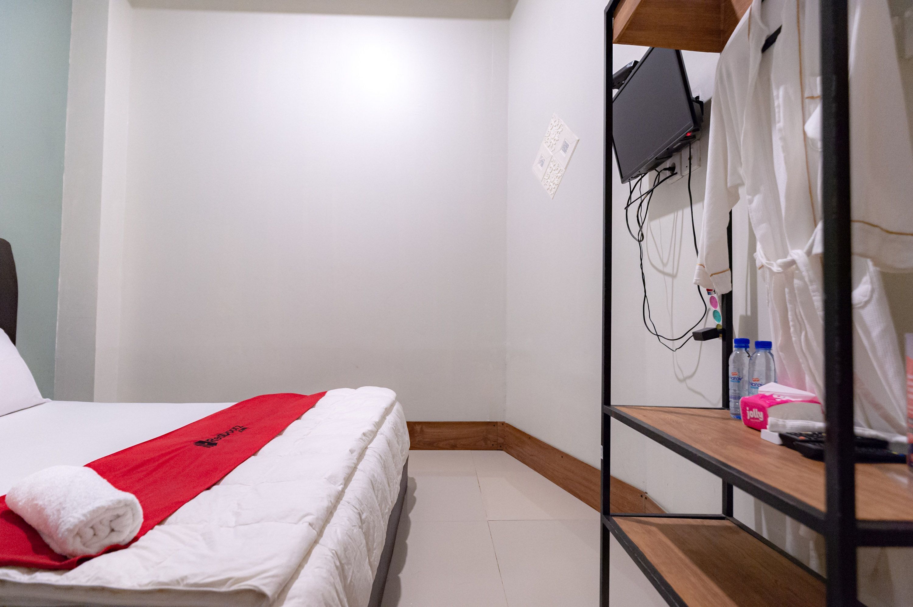 A hotel room with a bed, a red towel, a TV mounted on the wall, and a shelf with toiletries.