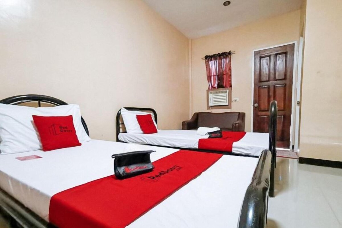 A hotel room with two beds, each with white bedding and red accents, a red blanket, and a red pillow. There is a chair and a wooden door in the room.