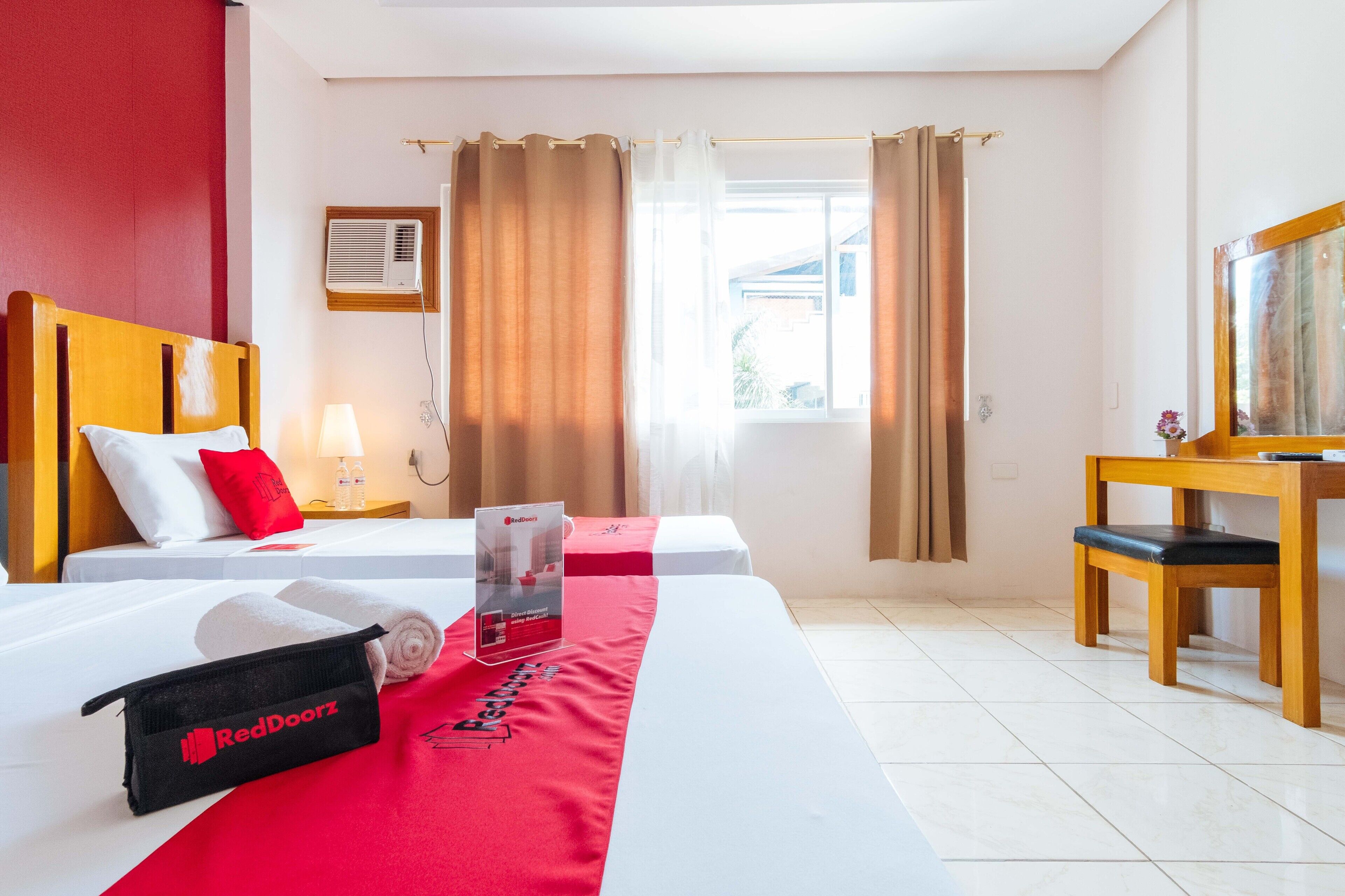 A hotel room with two beds, a desk, a chair, a lamp, a window with curtains, and a RedDoorz towel bag.