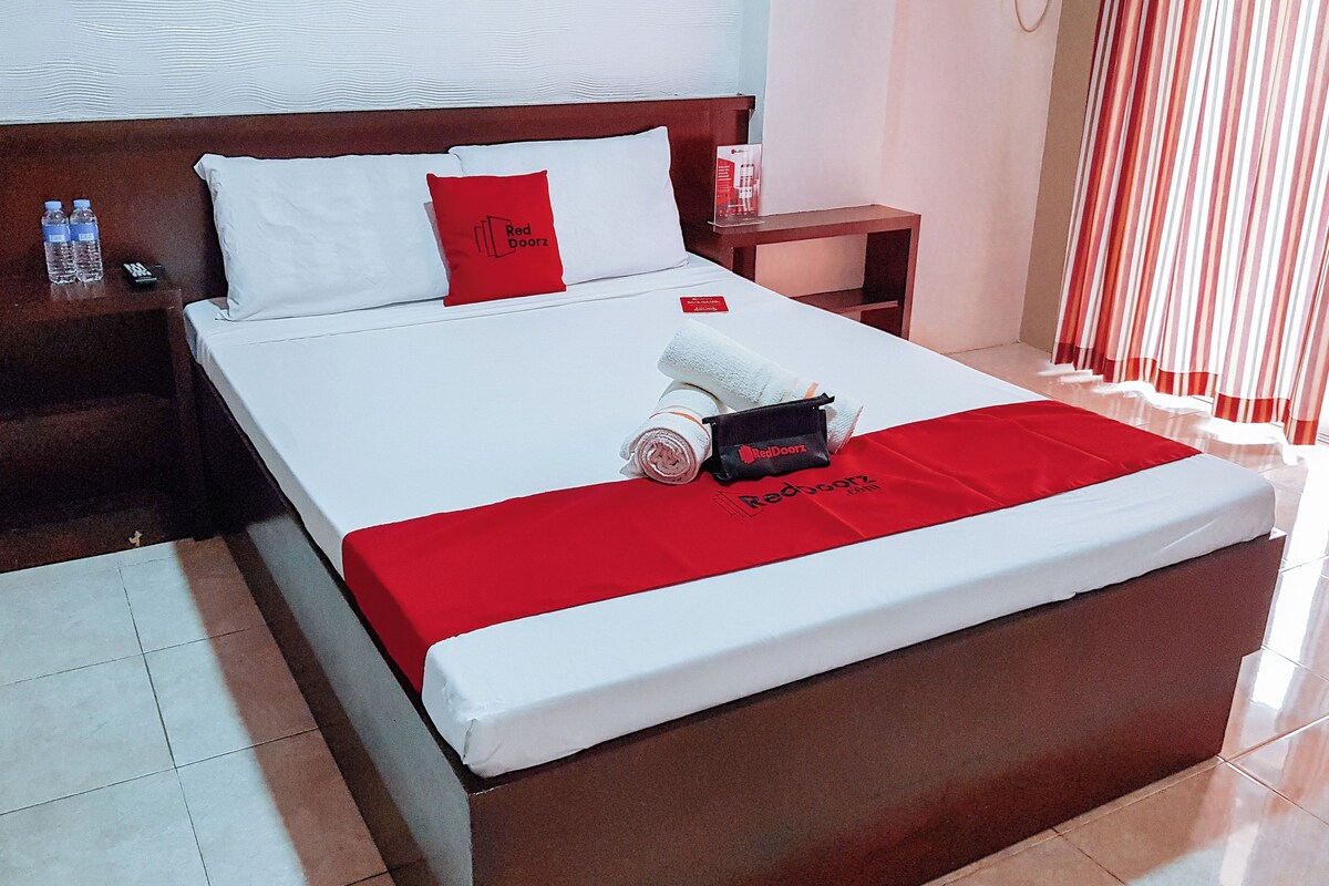 A neatly made bed with white and red bedding, a red pillow, and a towel with a RedDoorz logo. There is a small wooden nightstand with a remote control and water bottles beside the bed.