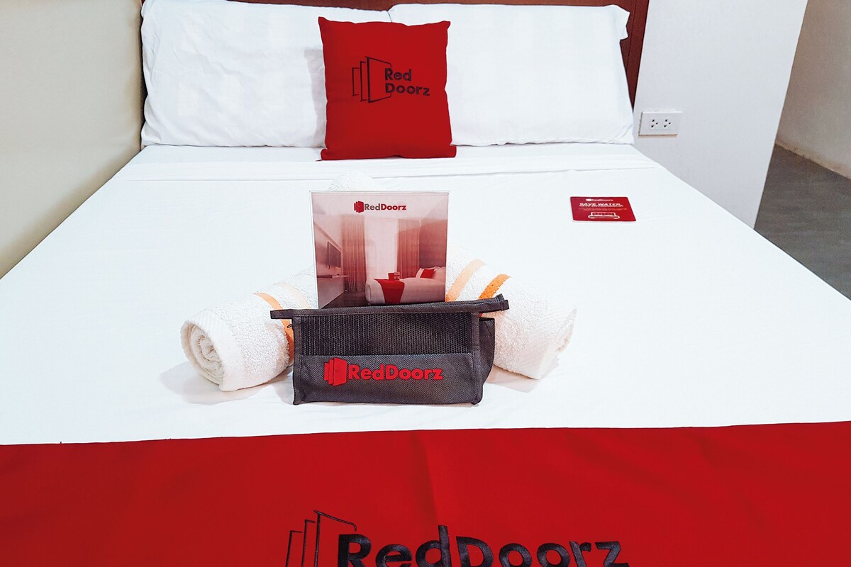 Ein ordentlich bezogenes Bett mit einem roten und weißen Kopfkissen, einer Tasche mit RedDoorz-Logo und einer RedDoorz-Broschüre.