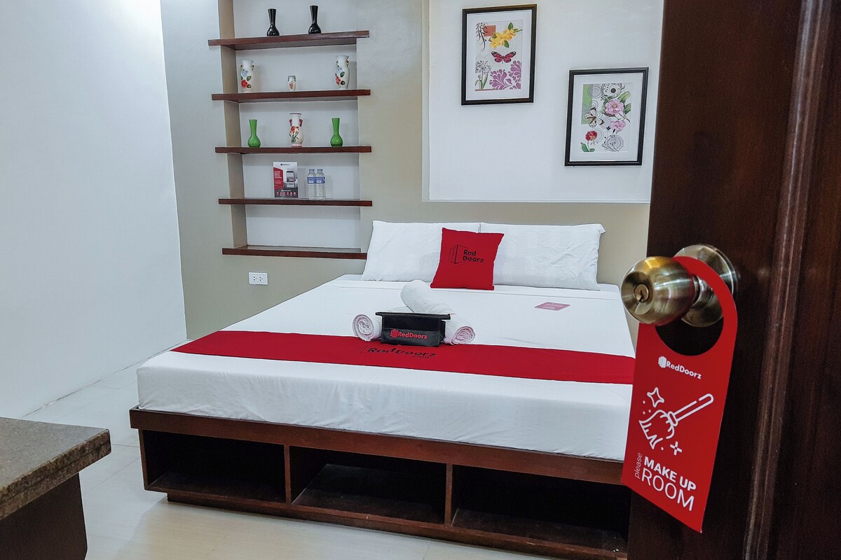 A hotel room with a bed, a red pillow, a red sign on the door, and a shelf with decorative items.
