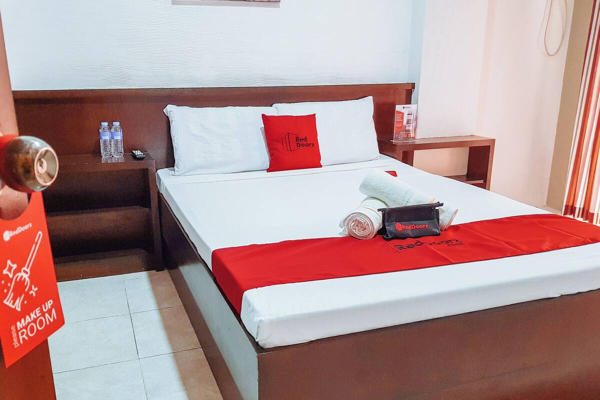 Hotellihuone, jossa on sänky, tyynyjä, yöpöytä, punainen pyyhe ja RedDoorz-kyltti.