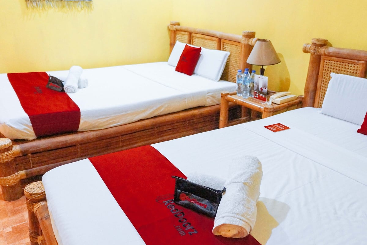 Two double beds with wooden headboards, white bedding, and red pillows. There is a nightstand with a lamp and a bottle of water between the beds.