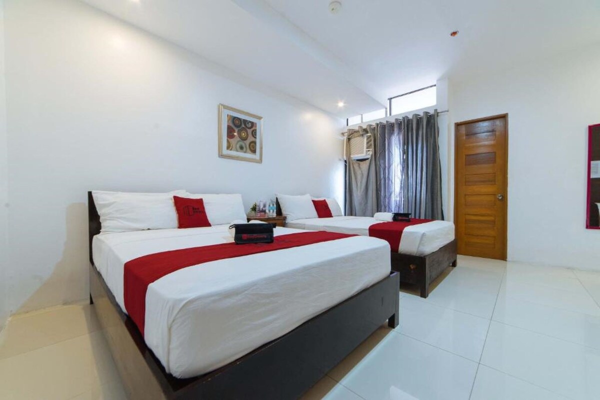 A hotel room with two beds, a wooden door, a window with curtains, and a wall-mounted air conditioning unit.
