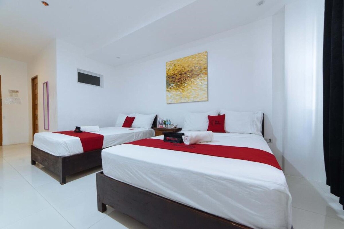 A hotel room with two beds, white bedding, red accents, a small painting on the wall, and a window with curtains.