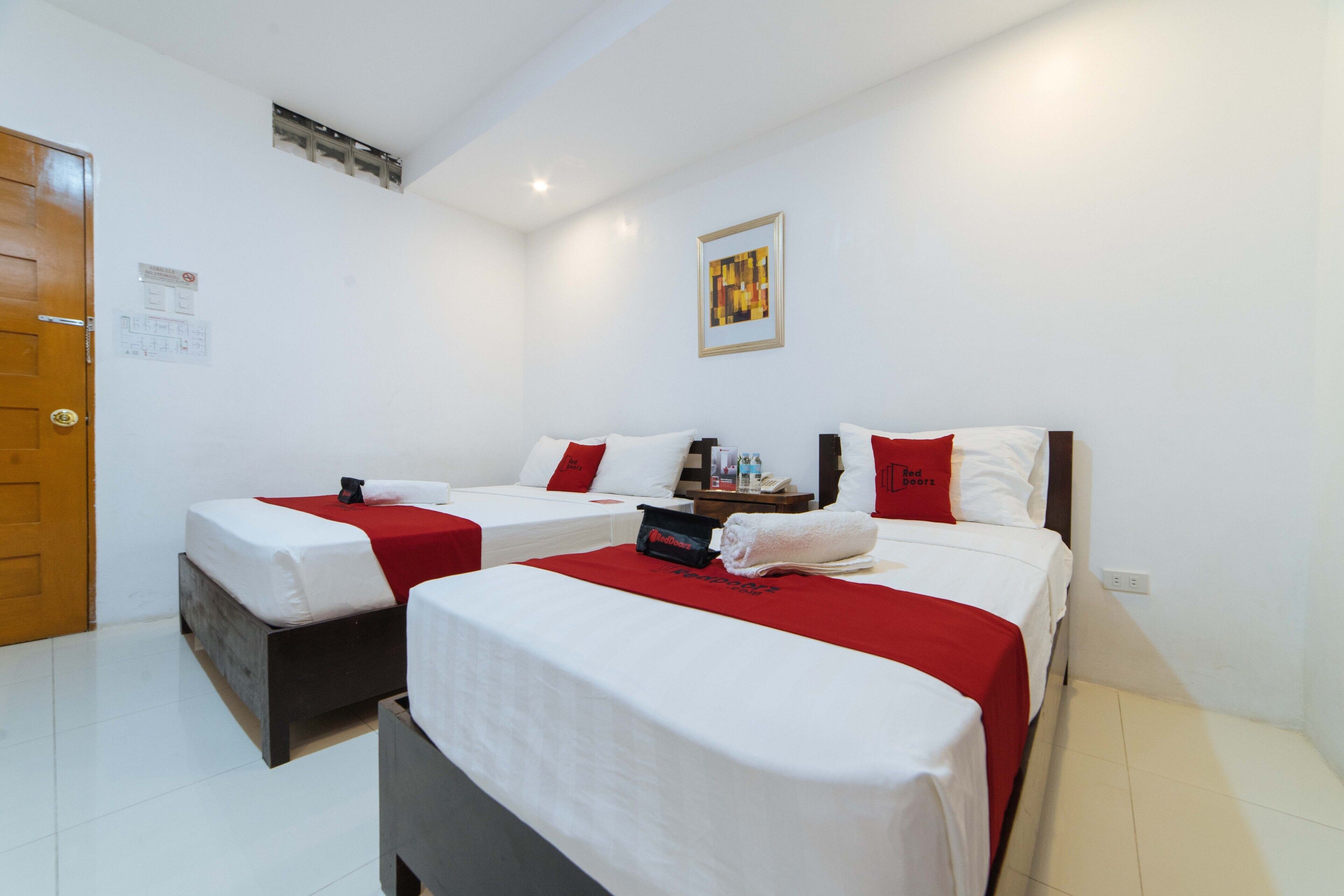 Una habitación de hotel con dos camas, cada una con ropa blanca y detalles rojos, una mesita de noche pequeña con una lámpara y una obra de arte enmarcada en la pared.