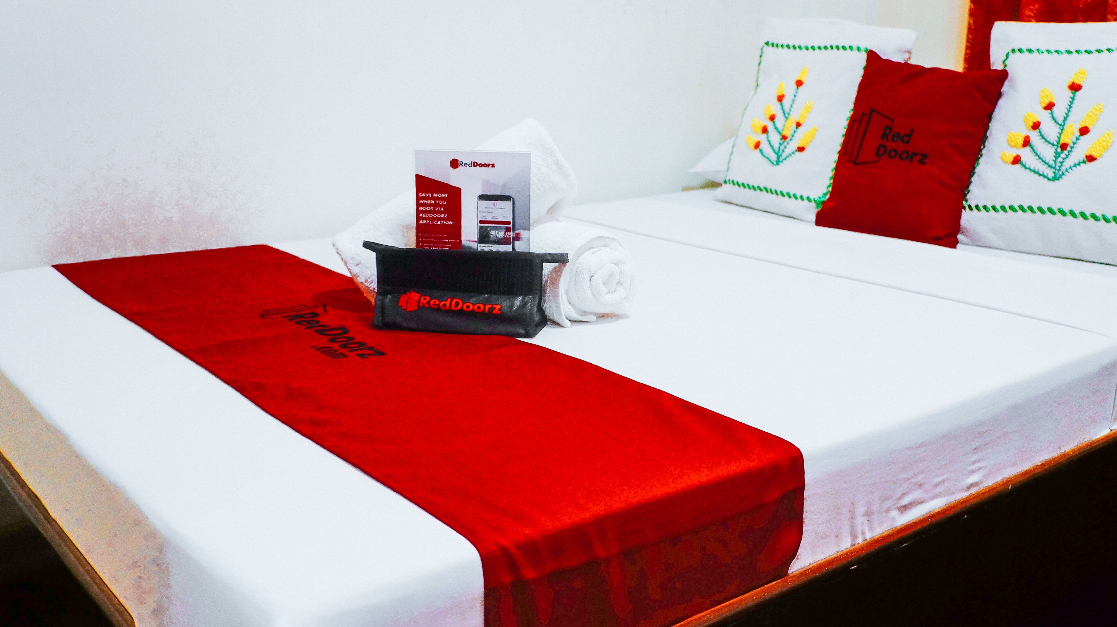 A neatly made bed with a red and white bedspread, a rolled white towel, and a RedDoorz branded phone charging station.