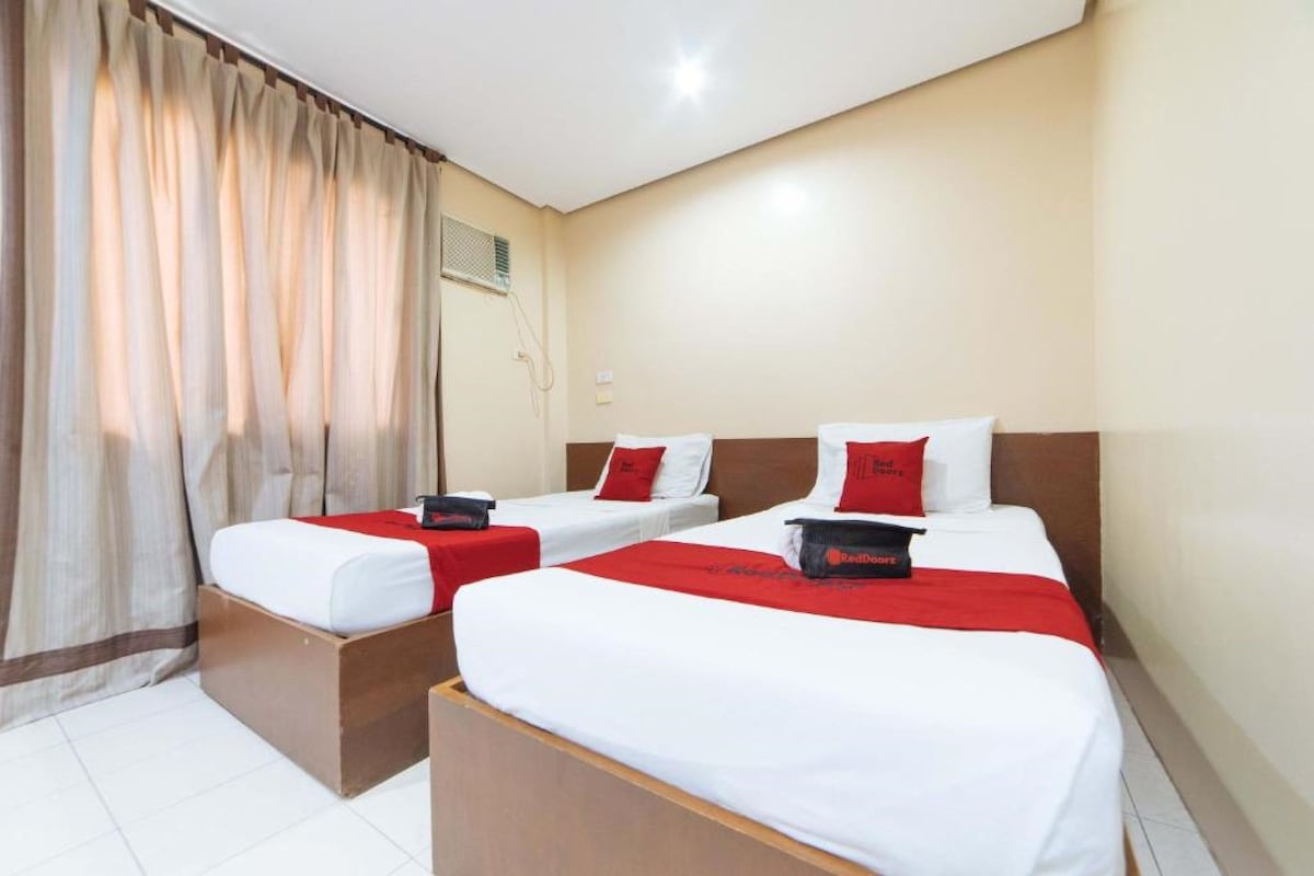 Habitación de hotel con dos camas, cada una con cabecero rojo, ropa blanca y una manta roja. Se ve una ventana con cortinas, un equipo de aire acondicionado montado en la pared y una mesita entre las camas.