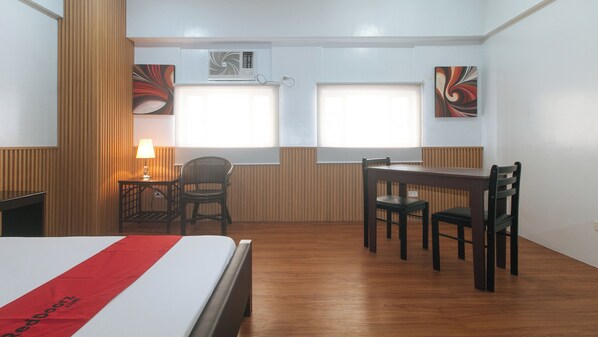 Habitación de hotel con cama, escritorio, dos sillas, lámpara y dos ventanas con persianas.