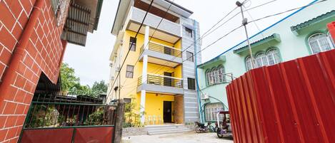 Un edificio residencial de varios pisos con fachada amarilla, un balcón en el segundo nivel y una cerca roja en primer plano.
