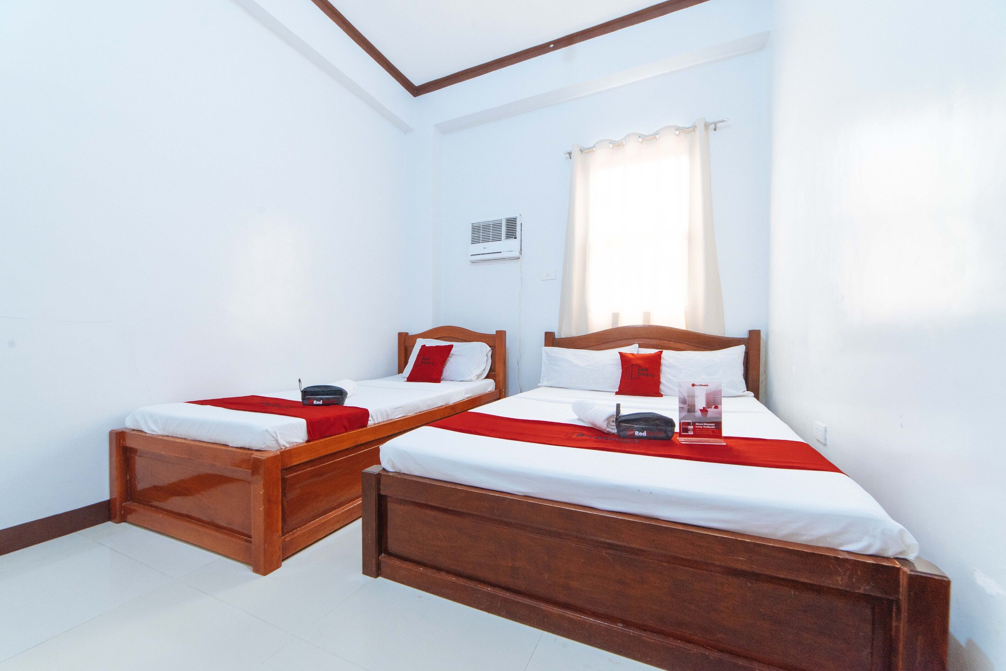 A room with two wooden beds, white bedding, red accents, a window with curtains, and an air conditioning unit.