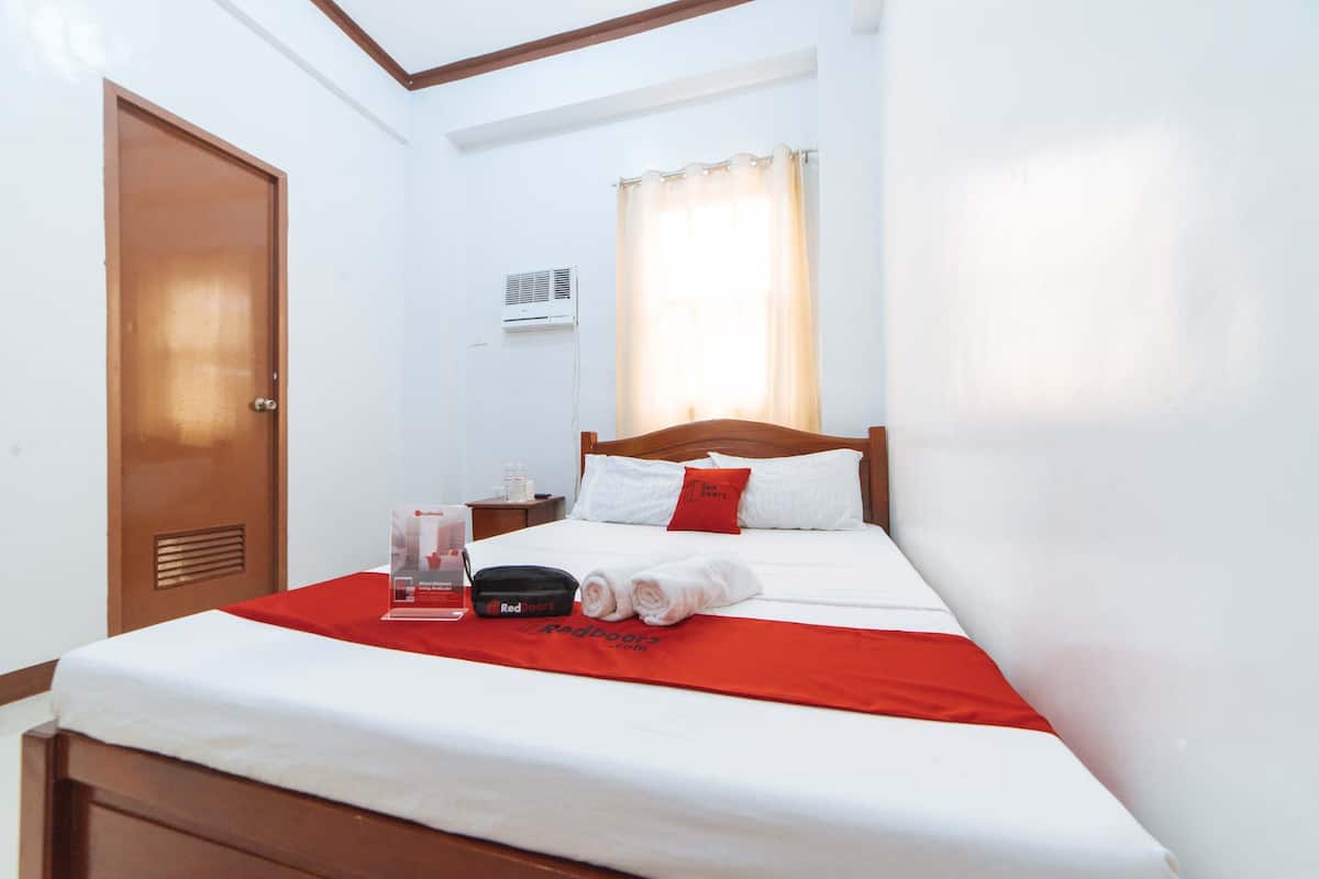 A hotel room with a bed, a nightstand, a red and white bedspread, a red pillow, a white pillow, a window with curtains, an air conditioning unit, and a door.