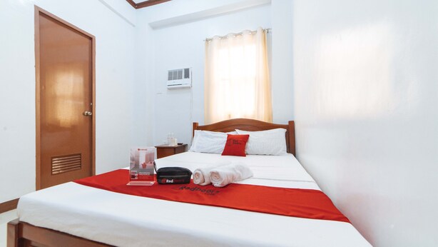 A neatly made bed with a red and white bedspread, a wooden headboard, and a pillow with a red logo. There is a nightstand with a lamp and a bottle of water. - Reddoorz @ D Maagma Street - Vaccinated Staff (Kalibo)