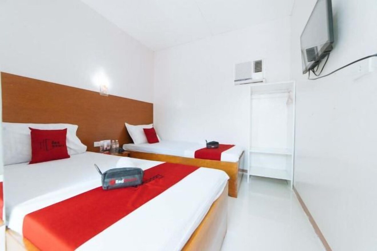 A hotel room with two beds, each with red and white bedding, a wooden headboard, a flat-screen TV mounted on the wall, and a window with a white blind.