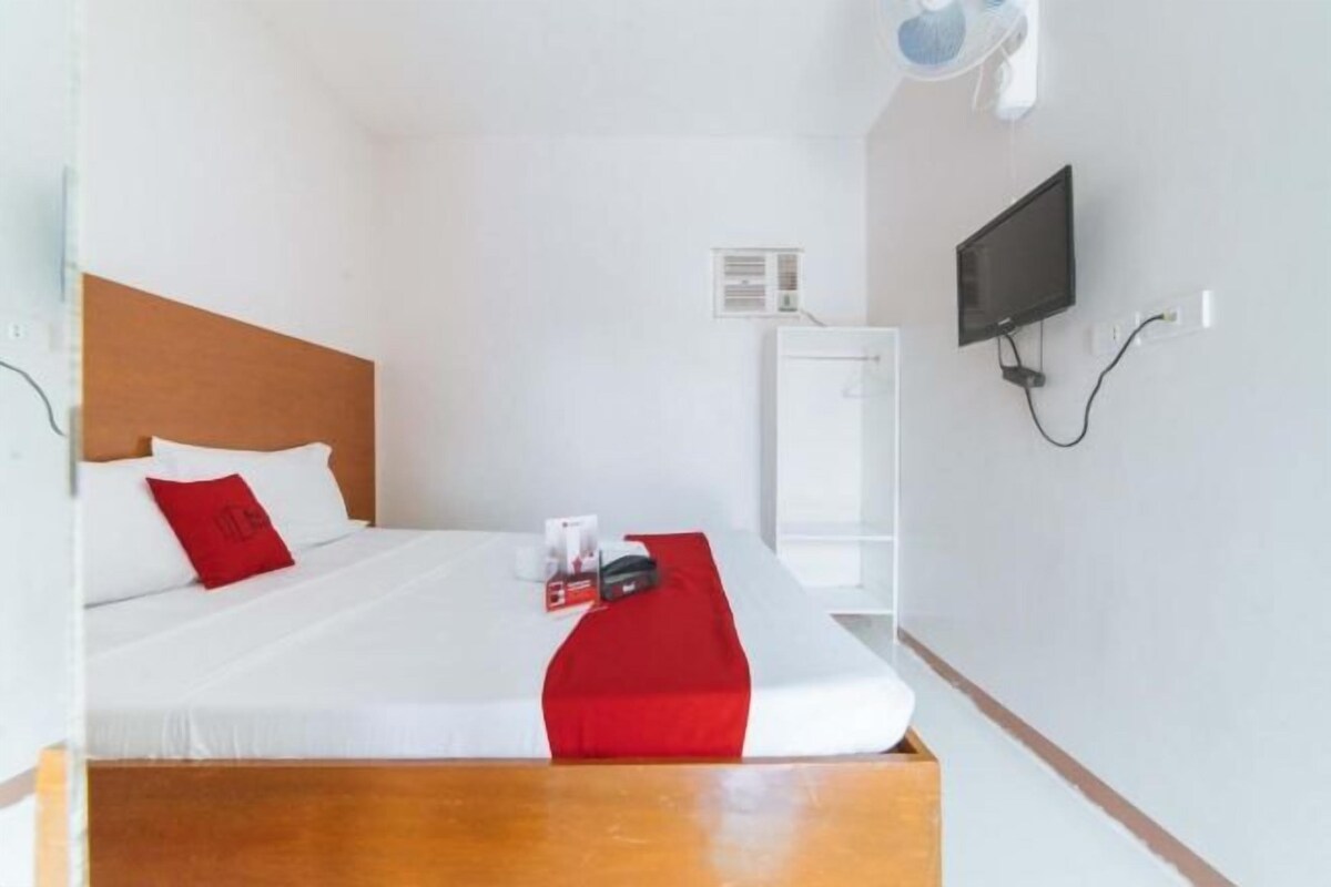 A hotel room with a single bed, a wooden headboard, a red accent pillow, a white bedspread, a red throw blanket, a bedside table, a TV mounted on the wall, and a white wardrobe.
