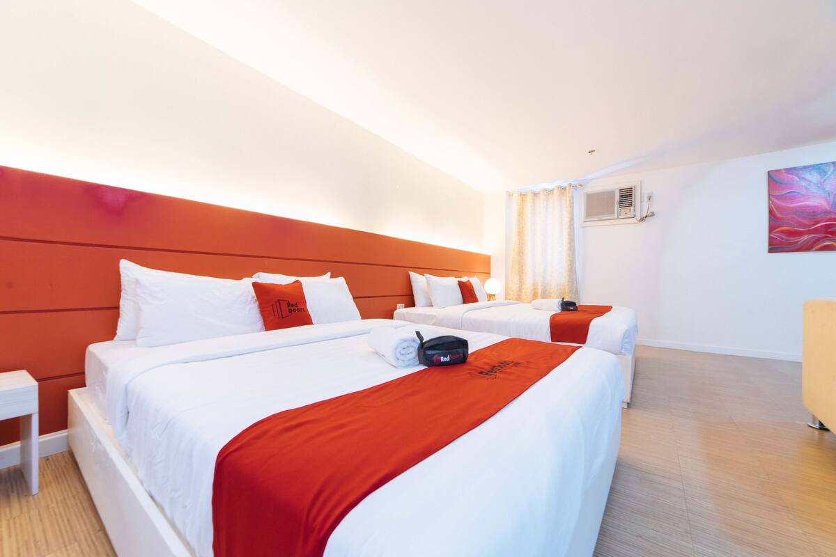 A hotel room with two beds, a red accent wall, white bedding, a bedside table, and a painting on the wall.