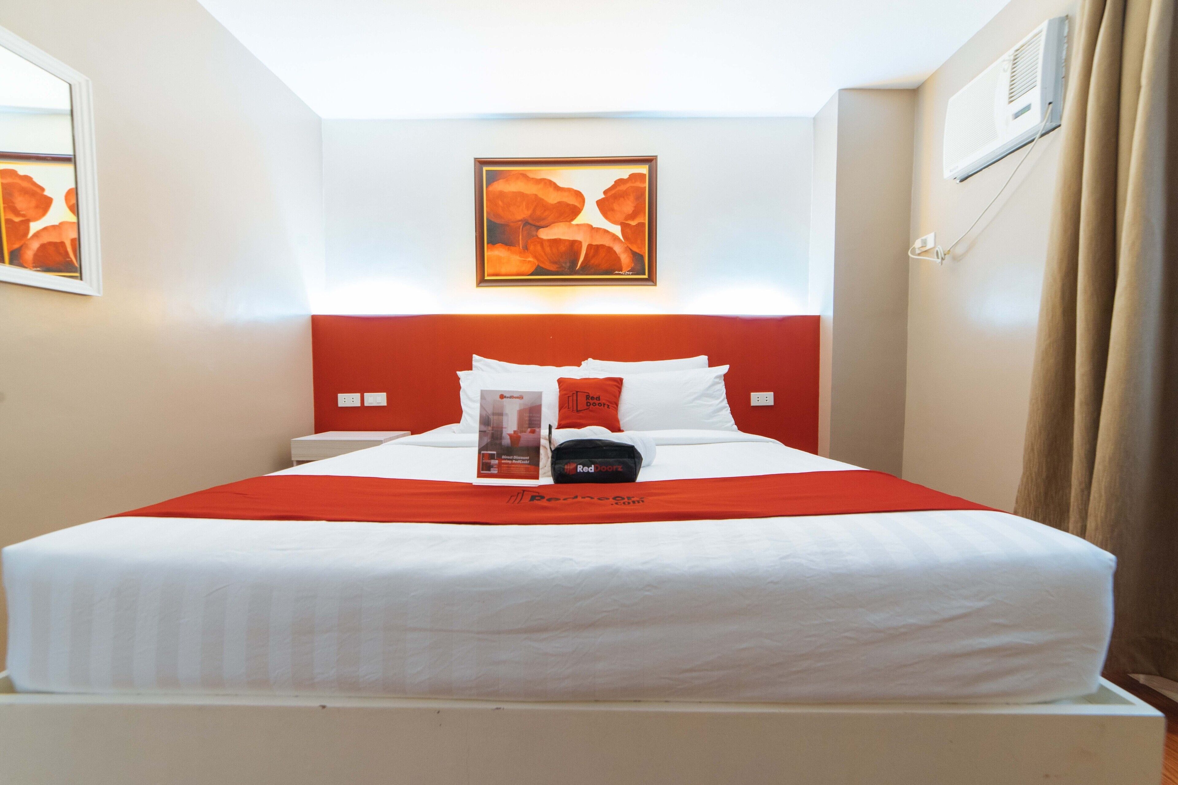A hotel room with a bed, a red headboard, a framed artwork above the bed, and a window with curtains.