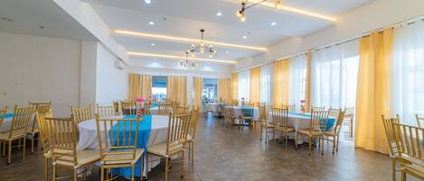 Un amplio comedor con varias mesas y sillas, luminarias modernas y ventanas grandes con cortinas.